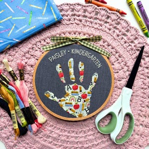 May include: A circular wooden embroidery hoop containing dark gray fabric with a handprint applique featuring a pattern of apples, books, and rainbows. The text PAISLEY KINDERGARTEN is embroidered in white above the handprint, topped with a green gingham ribbon.