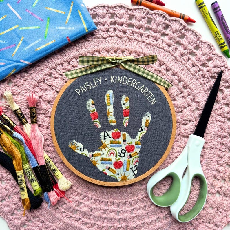 May include: A circular wooden embroidery hoop containing dark gray fabric with a handprint applique featuring a pattern of apples, books, and rainbows. The text PAISLEY KINDERGARTEN is embroidered in white above the handprint, topped with a green gingham ribbon.