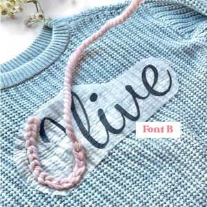May include: A light blue knitted sweater with the word "olive" written in black on a white background. A pink braided rope is wrapped around the word.