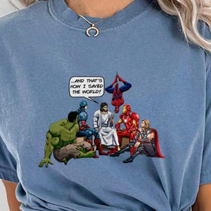 May include: Blue jean colored t-shirt featuring a comic-style illustration. The graphic depicts several superheroes and a central figure, with a speech bubble stating, "...AND THAT'S HOW I SAVED THE WORLD!"