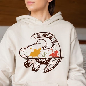 May include: A cream-colored hoodie with a graphic of the Lion King's Simba, Nala, and Timon in a stylized outline of the Pride Rock symbol. The graphic is in shades of brown, red, yellow, and green.