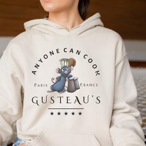 May include: A white hoodie with a graphic of a rat chef wearing a toque and holding a salt shaker and pepper shaker. The text on the hoodie reads 'Anyone Can Cook', 'Paris', 'France', 'Gusteau's', and five stars.