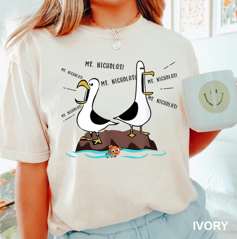 K&ouml;nnte beinhalten: Elfenbeinfarbenes T-Shirt mit einer Cartoon-Illustration von zwei M&ouml;wen mit dem Text "MS. NICHOLAS!". Die M&ouml;wen stehen auf einem Felsen &uuml;ber dem Wasser, darunter ein kleiner Fisch. Eine wei&szlig;e Tasse mit einem Smiley ist ebenfalls sichtbar.