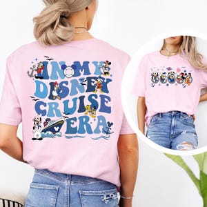 May include: A light pink t-shirt featuring a large graphic on the back with the text IN MY DISNEY CRUISE ERA in blue wavy letters, surrounded by illustrations of Disney characters in nautical outfits, a cruise ship, and anchors.