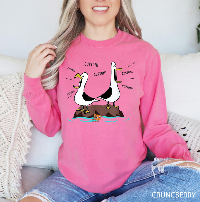K&ouml;nnte beinhalten: Ein pinkfarbenes Langarm-Sweatshirt mit einer Cartoon-Illustration von zwei M&ouml;wen auf einem Felsen, wobei das Wort "CUSTOM" aus ihren Schn&auml;beln kommt. Ein kleiner Fisch befindet sich im Wasser darunter. Das Wort "CRUNCBERRY" steht unten.