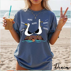 K&ouml;nnte beinhalten: Denimblaues T-Shirt mit einem Cartoon-Design von zwei M&ouml;wen auf einem Felsen, mit dem Wort "MAMA" in Sprechblasen. Ein kleiner oranger Fisch schwimmt darunter. Das Wort "Denim" ist unten rechts in Kursivschrift geschrieben.