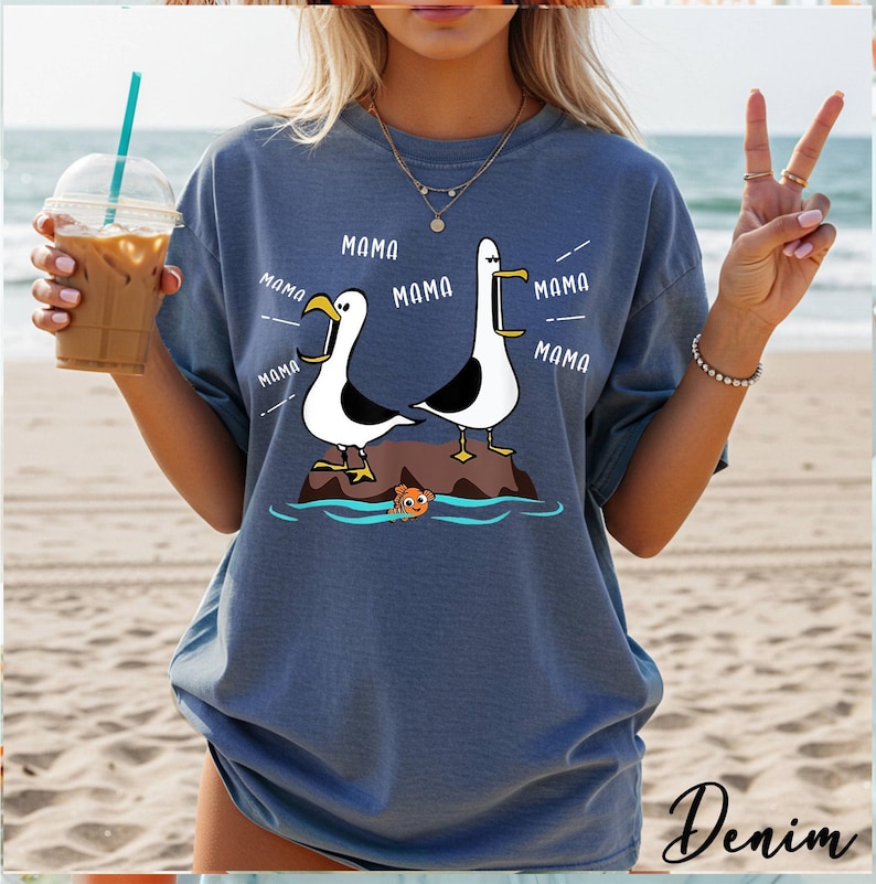 K&ouml;nnte beinhalten: Denimblaues T-Shirt mit einem Cartoon-Design von zwei M&ouml;wen auf einem Felsen, mit dem Wort "MAMA" in Sprechblasen. Ein kleiner oranger Fisch schwimmt darunter. Das Wort "Denim" ist unten rechts in Kursivschrift geschrieben.