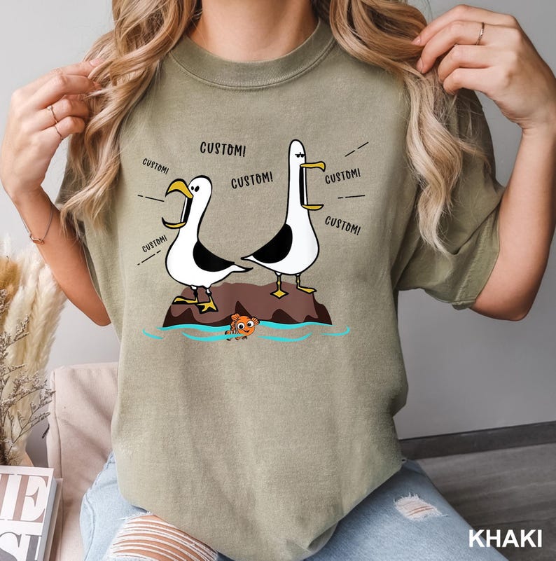 K&ouml;nnte beinhalten: Khaki-farbenes T-Shirt mit einer Cartoon-Illustration von zwei M&ouml;wen auf einem Felsen, mit einem Fisch im Wasser. &Uuml;ber den M&ouml;wen steht das Wort "CUSTOM!". Das T-Shirt hat eine lockere Passform.