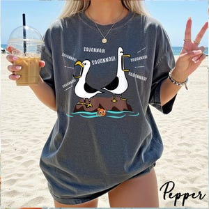 K&ouml;nnte beinhalten: Dunkelgraues T-Shirt mit einer Cartoon-Illustration von zwei M&ouml;wen auf einem Felsen, mit dem Text "SAVANNAH!" wiederholt. Ein kleiner orangefarbener Fisch ist im Wasser zu sehen. Das Shirt hat eine lockere Passform.