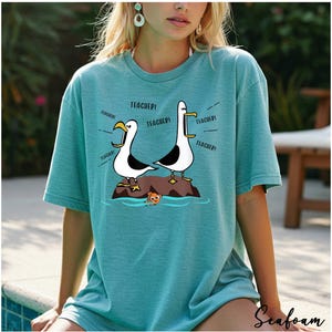 K&ouml;nnte beinhalten: T&uuml;rkisfarbenes T-Shirt mit einem Cartoon-Design von zwei M&ouml;wen auf einem Felsen, die "TEACHER!" schreien. Ein kleiner orangefarbener Fisch schwimmt darunter. Das T-Shirt hat kurze &Auml;rmel und eine lockere Passform.