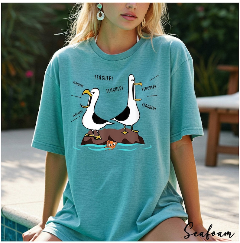 K&ouml;nnte beinhalten: T&uuml;rkisfarbenes T-Shirt mit einem Cartoon-Design von zwei M&ouml;wen auf einem Felsen, die "TEACHER!" schreien. Ein kleiner orangefarbener Fisch schwimmt darunter. Das T-Shirt hat kurze &Auml;rmel und eine lockere Passform.