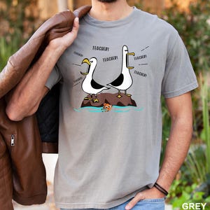 K&ouml;nnte beinhalten: Graues T-Shirt mit einer Cartoon-Illustration von zwei M&ouml;wen, die "TEACHER!" schreien. Die M&ouml;wen stehen auf einem Felsen, mit einem kleinen Fisch im Wasser. Das T-Shirt hat eine einfarbige graue Farbe.