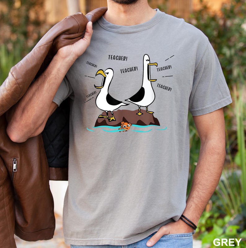 K&ouml;nnte beinhalten: Graues T-Shirt mit einer Cartoon-Illustration von zwei M&ouml;wen, die "TEACHER!" schreien. Die M&ouml;wen stehen auf einem Felsen, mit einem kleinen Fisch im Wasser. Das T-Shirt hat eine einfarbige graue Farbe.