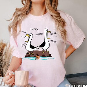 K&ouml;nnte beinhalten: Hellrosa T-Shirt mit einem Cartoon-Design von zwei M&ouml;wen, die "Teacher!" schreien. Die M&ouml;wen stehen auf einer kleinen Insel mit einem Cartoon-Fisch. Das Wort "BLOSSOM" steht unten rechts.