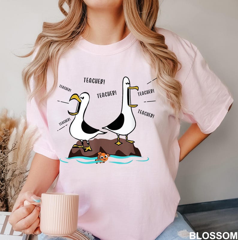 K&ouml;nnte beinhalten: Hellrosa T-Shirt mit einem Cartoon-Design von zwei M&ouml;wen, die "Teacher!" schreien. Die M&ouml;wen stehen auf einer kleinen Insel mit einem Cartoon-Fisch. Das Wort "BLOSSOM" steht unten rechts.