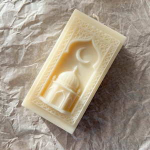 K&ouml;nnte beinhalten: Elfenbeinfarbene rechteckige Kerze mit detailliertem Reliefdesign. Das Design zeigt eine Moschee mit Kuppel und Mondsichel, umrahmt von einem verzierten Rand. Die Kerze ist unparf&uuml;miert und hat ein nat&uuml;rliches, handgemachtes Aussehen.