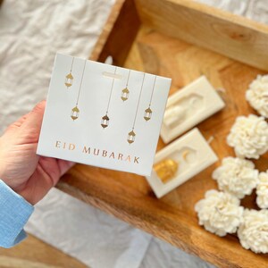 K&ouml;nnte beinhalten: Eine wei&szlig;e Karte mit goldenen Laternenillustrationen und dem Text "EID MUBARAK" wird in einer Hand gehalten. Die Karte liegt auf einem Holztablett mit wei&szlig;en Blumendekorationen und zwei wei&szlig;en rechteckigen Seifen mit goldenen Akzenten.