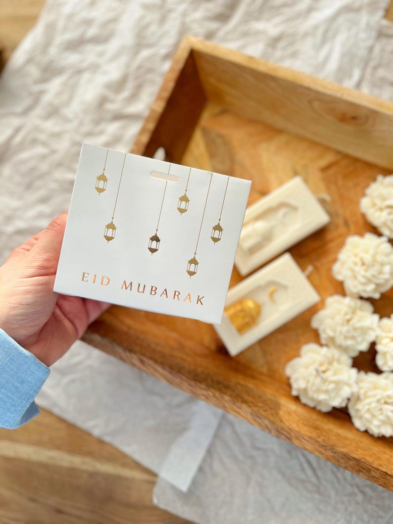 K&ouml;nnte beinhalten: Eine wei&szlig;e Karte mit goldenen Laternenillustrationen und dem Text "EID MUBARAK" wird in einer Hand gehalten. Die Karte liegt auf einem Holztablett mit wei&szlig;en Blumendekorationen und zwei wei&szlig;en rechteckigen Seifen mit goldenen Akzenten.