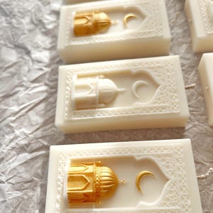 K&ouml;nnte beinhalten: Elfenbeinfarbene rechteckige Kerzen mit goldenen Moschee- und Mondsichel-Designs. Die Kerzen haben komplizierte, gepr&auml;gte Muster an den R&auml;ndern. Die Moscheedome sind goldfarben bemalt, und die Halbmonde sind ebenfalls goldfarben.