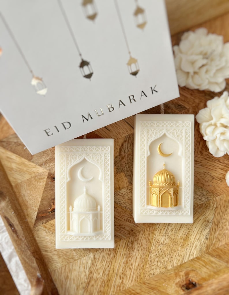 K&ouml;nnte beinhalten: Zwei rechteckige, cremefarbene Seifen mit Moschee- und Mond-Designs. Eine Seife zeigt eine goldene Moschee. Eine Gru&szlig;karte mit den Worten "EID MUBARAK" und Illustrationen von h&auml;ngenden Laternen im Hintergrund.