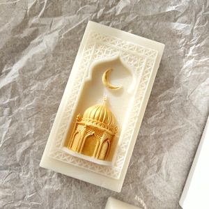 K&ouml;nnte beinhalten: Elfenbeinfarbene Kerze mit einem goldenen Moschee- und Mond-Design. Die rechteckige Kerze hat einen gepr&auml;gten Rahmen. Die Moschee und der Mond sind erhaben und goldfarben bemalt, was einen dekorativen Akzent setzt.