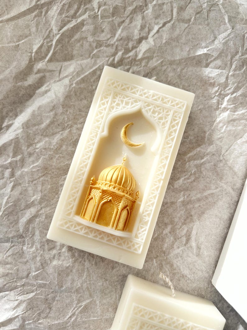 K&ouml;nnte beinhalten: Elfenbeinfarbene Kerze mit einem goldenen Moschee- und Mond-Design. Die rechteckige Kerze hat einen gepr&auml;gten Rahmen. Die Moschee und der Mond sind erhaben und goldfarben bemalt, was einen dekorativen Akzent setzt.