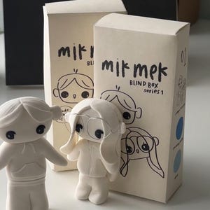 Puede incluir: Dos figuritas de cerámica blanca de personas con rasgos simples, de pie frente a dos cajas con el texto "mik mek blind box series 1". Las cajas son beige y tienen un diseño simple.