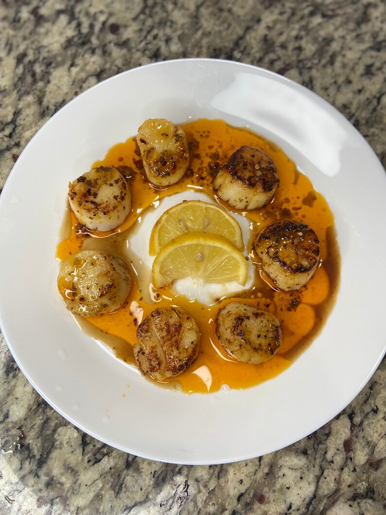 May include: A white plate with six seared scallops, lemon wedges, and a bright orange sauce.