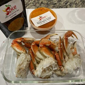 May include: A glass baking dish filled with cooked crab legs. The dish is on a white surface. There is a small container of red sauce and a bag of crab boil seasoning next to the dish.