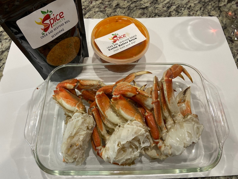 May include: A glass baking dish filled with cooked crab legs. The dish is on a white surface. There is a small container of red sauce and a bag of crab boil seasoning next to the dish.