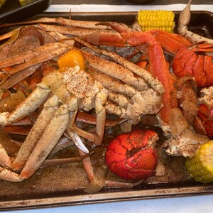 May include: A metal baking sheet filled with a seafood boil. The sheet contains crab legs, lobster tails, corn on the cob, and a yellow pepper. The seafood is seasoned with spices and appears to be cooked.