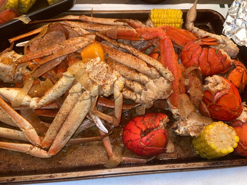 May include: A metal baking sheet filled with a seafood boil. The sheet contains crab legs, lobster tails, corn on the cob, and a yellow pepper. The seafood is seasoned with spices and appears to be cooked.