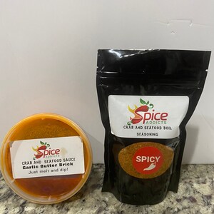May include: Two containers of seafood seasoning. One is a red plastic container with a label that reads "Spice Addicts Crab and Seafood Sauce Garlic Butter Brick Just melt and dip!" The other is a black plastic bag with a label that reads "Spice Addicts Crab and Seafood Boil Seasoning" with the word "Spicy" in red.