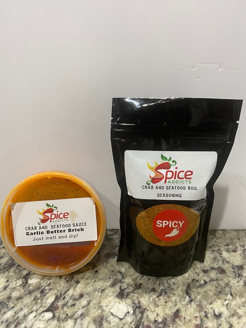 May include: Two containers of seafood seasoning. One is a red plastic container with a label that reads "Spice Addicts Crab and Seafood Sauce Garlic Butter Brick Just melt and dip!" The other is a black plastic bag with a label that reads "Spice Addicts Crab and Seafood Boil Seasoning" with the word "Spicy" in red.