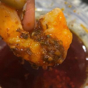May include: A close-up of a shrimp dipped in a red sauce with herbs and spices. The shrimp is being held by a finger and is partially submerged in the sauce.