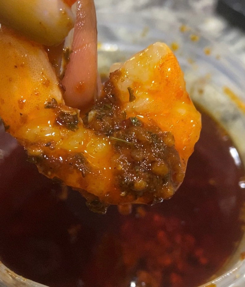 May include: A close-up of a shrimp dipped in a red sauce with herbs and spices. The shrimp is being held by a finger and is partially submerged in the sauce.