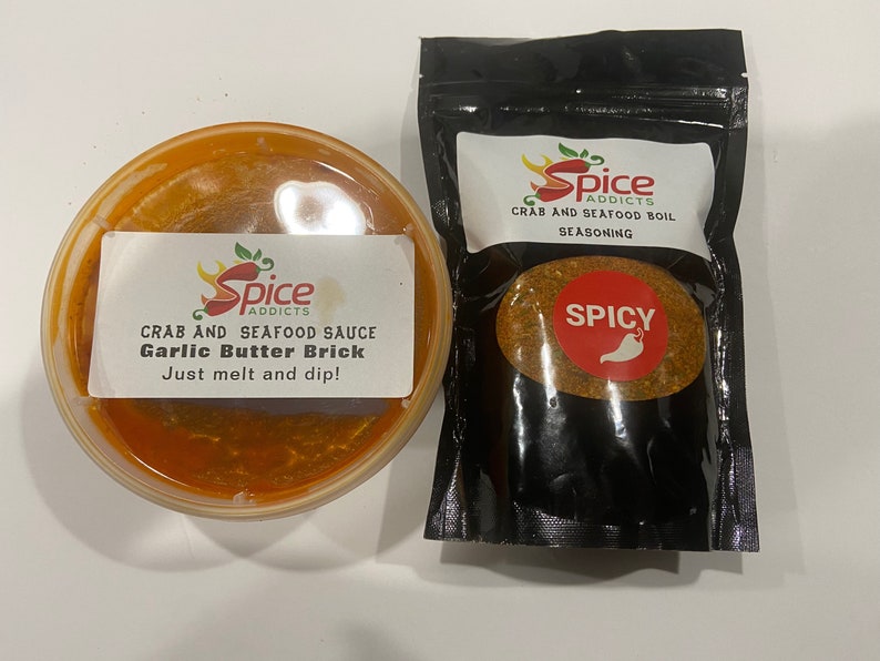 May include: Two containers of crab and seafood seasoning. One is a red sauce in a clear plastic container with a label that reads "Spice Addicts Crab and Seafood Sauce Garlic Butter Brick Just melt and dip!" The other is a black bag with a label that reads "Spice Addicts Crab and Seafood Boil Seasoning" with a red circle that says "Spicy" and a red chili pepper.
