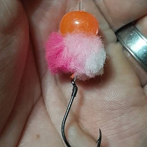 May include: A pink, white, and orange fishing fly with a black hook. The fly is made with a fluffy material and a small orange bead.