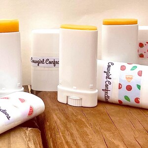 May include: A collection of white plastic lip balm tubes with yellow caps. Some tubes feature labels with the text Cowgirl Confections and various patterns including pumpkins, strawberries, and cherries. The tubes are arranged in an upright and horizontal group.