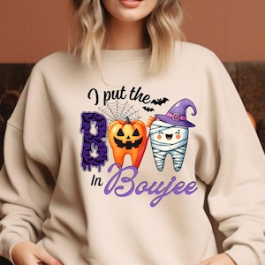 Può includere: Felpa beige con grafica a tema Halloween. La grafica presenta tre denti dei cartoni animati con la scritta "I put the Boo in Boujee". Un dente è bendato, un altro è una zucca e il terzo è viola con una ragnatela.