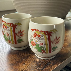 May include: Two white ceramic teacups with a floral and bamboo design. The cups have red, yellow, green, and gold accents.