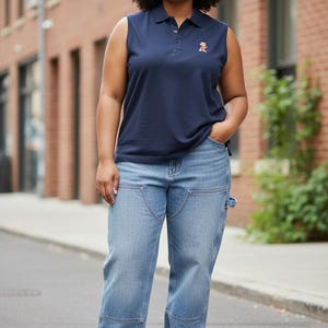 May include: A sleeveless navy blue polo shirt with a small embroidered design and light blue jeans. The jeans have a unique pocket design and are paired with white sneakers. The person is standing on a city street.
