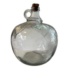 May include: A clear glass jug with a round body, a handle, and a cork stopper. The jug features leaf designs etched on the surface. The jug is empty and appears to be vintage.
