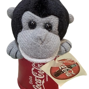 May include: A plush toy monkey with a gray face and black fur on top of its head. The monkey is holding a red Coca-Cola can with the Coca-Cola logo. A tag with the Coca-Cola logo is attached to the can.