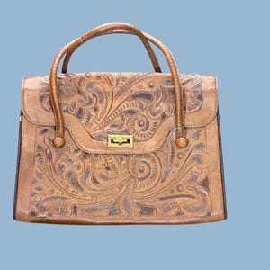 Pode incluir: Uma mala de mão de couro castanho com um design floral em relevo. A mala tem duas alças e um fecho dourado. A mala é retangular e tem um fecho de aba.