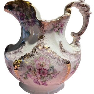 Puede incluir: Una jarra de porcelana blanca con un diseño floral y detalles dorados. La jarra tiene una boca ancha y un asa con un diseño de rollo decorativo.