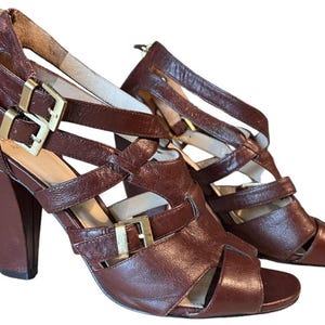 May include: Brown leather strappy sandals with gold-tone buckles and a chunky heel. The open-toe sandals feature multiple straps and a zipper closure at the back. The heel measures approximately 4 inches.