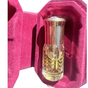 May include: A small, clear glass perfume bottle with a gold-colored metallic cap, featuring etched floral vine patterns on the sides. The bottle is nestled inside a deep magenta-colored velvet-lined case with a hinged opening.