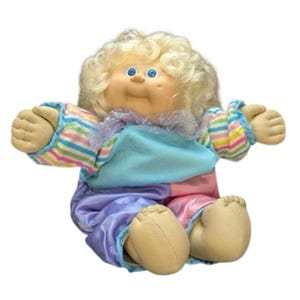 以下が含まれることがあります： ブロンドの巻き毛、青い目、バラ色の肌をしたヴィンテージ人形。人形は、水色のトップス、ピンクと紫のパンツ、ストライプの袖が付いたカラフルな衣装を着ています。人形は腕を広げて座っています。