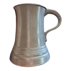 May include: A pewter-coloured beer stein with a handle. The mug has a wide base that tapers upwards to a narrower top. The handle is curved and attached to the side of the mug. The stein has a simple, classic design.